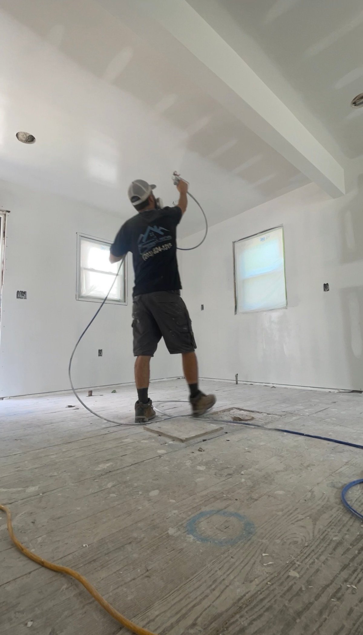 Property Revival crew member in branded shirt spraying drywall compound on a ceiling in an unfinished interior room with windows