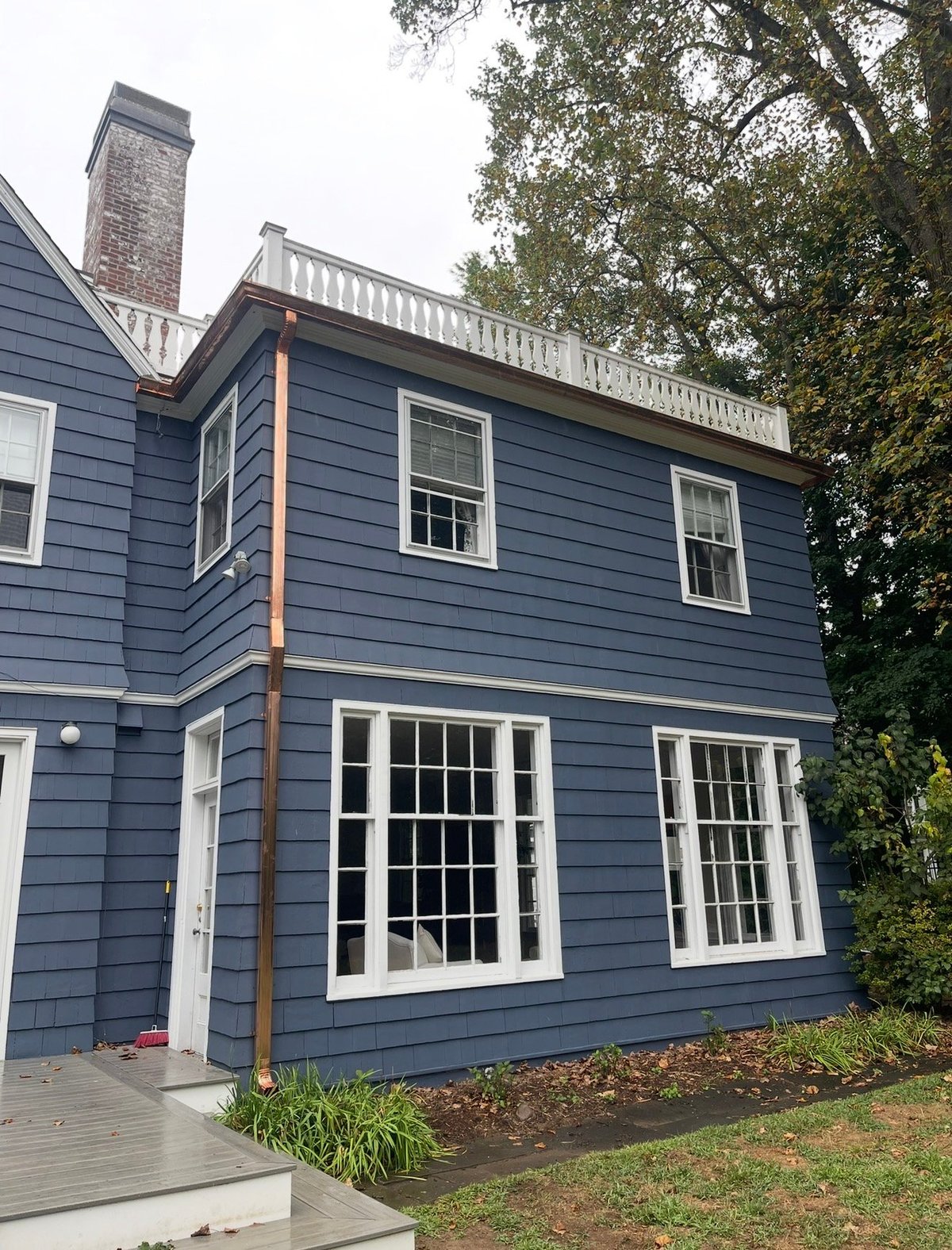 Two-story blue house with white-trimmed double-hung windows, brick chimney, white entry door, and a small upper balcony