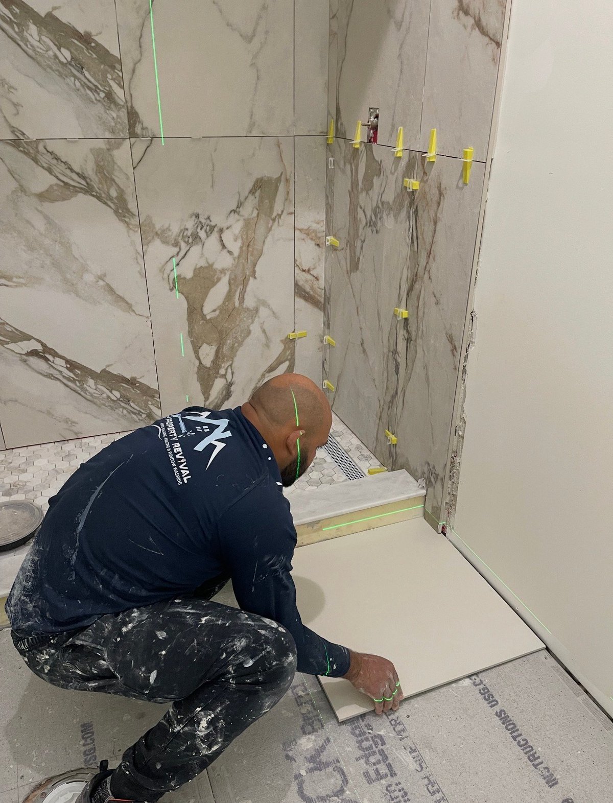 Worker installing marble tiles on a bathroom wall with yellow spacer clips and a level tool for precise alignment