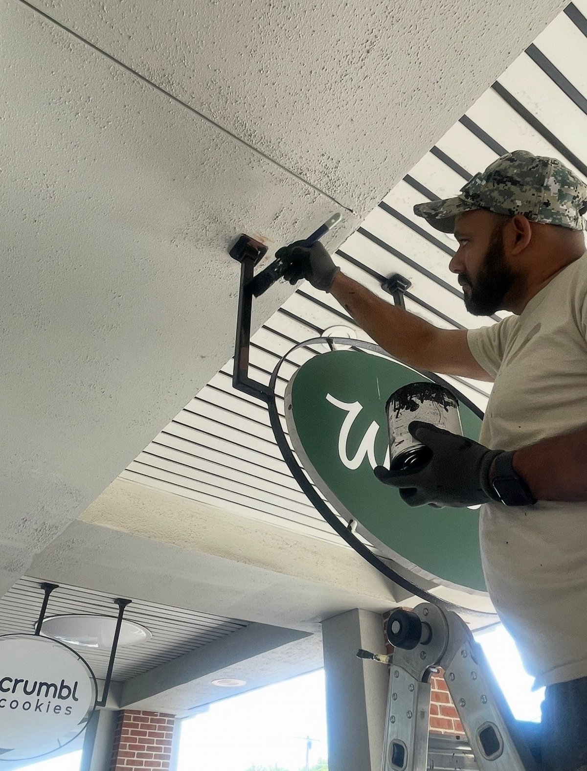 Worker wearing a cap and gloves installing a green circular sign on an outdoor storefront ceiling