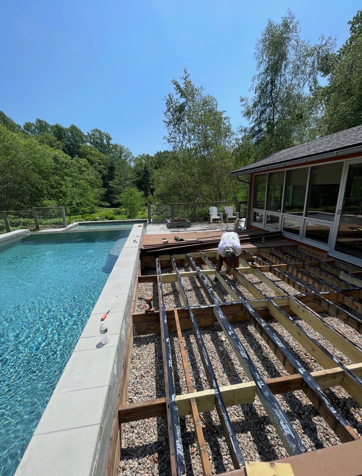 Backyard pool under construction with exposed deck framing beside a turquoise pool and a modern home among trees