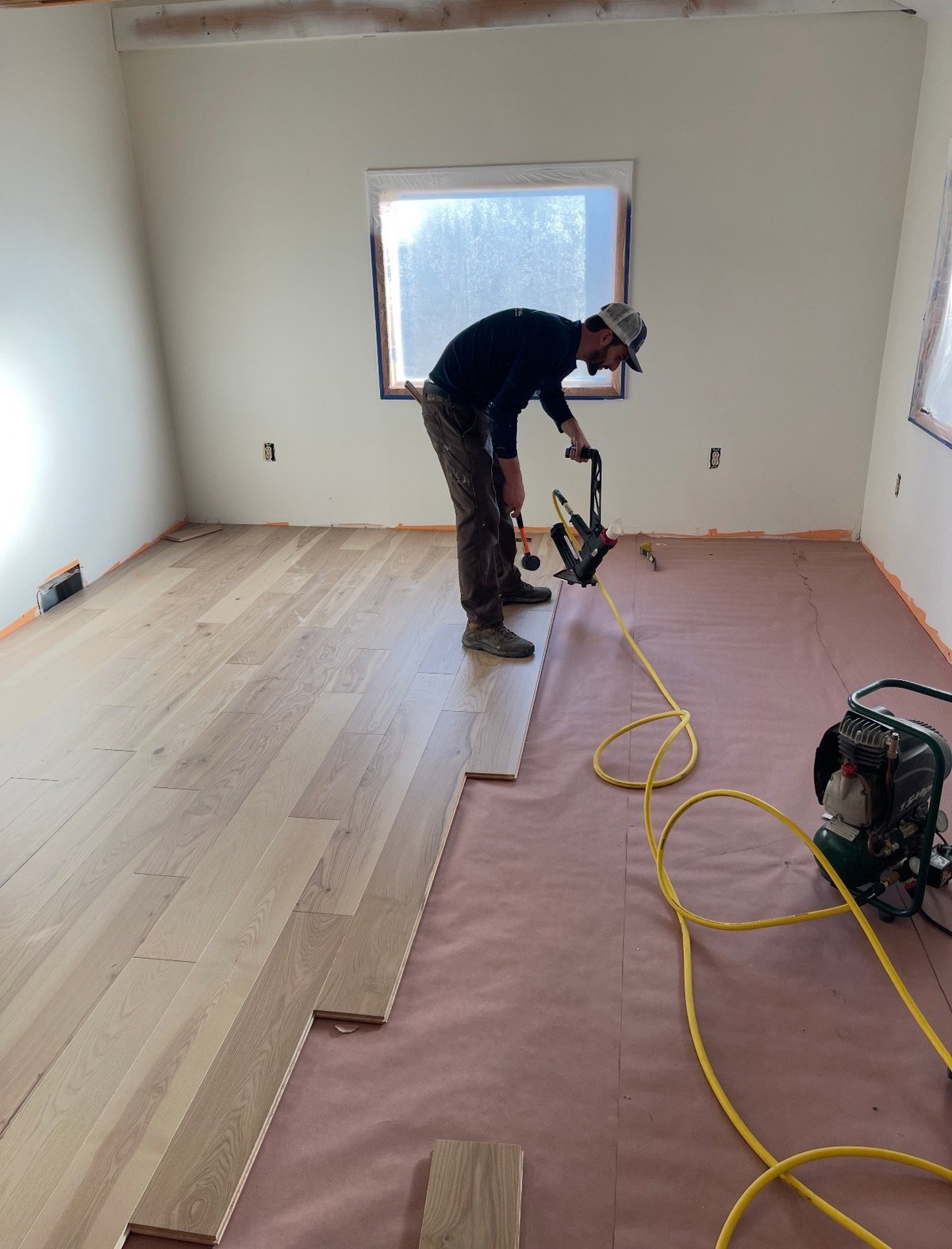 Property Revival crew member installing hardwood flooring with a nail gun, pink protective underlayment, and yellow air hose in an empty room with a window
